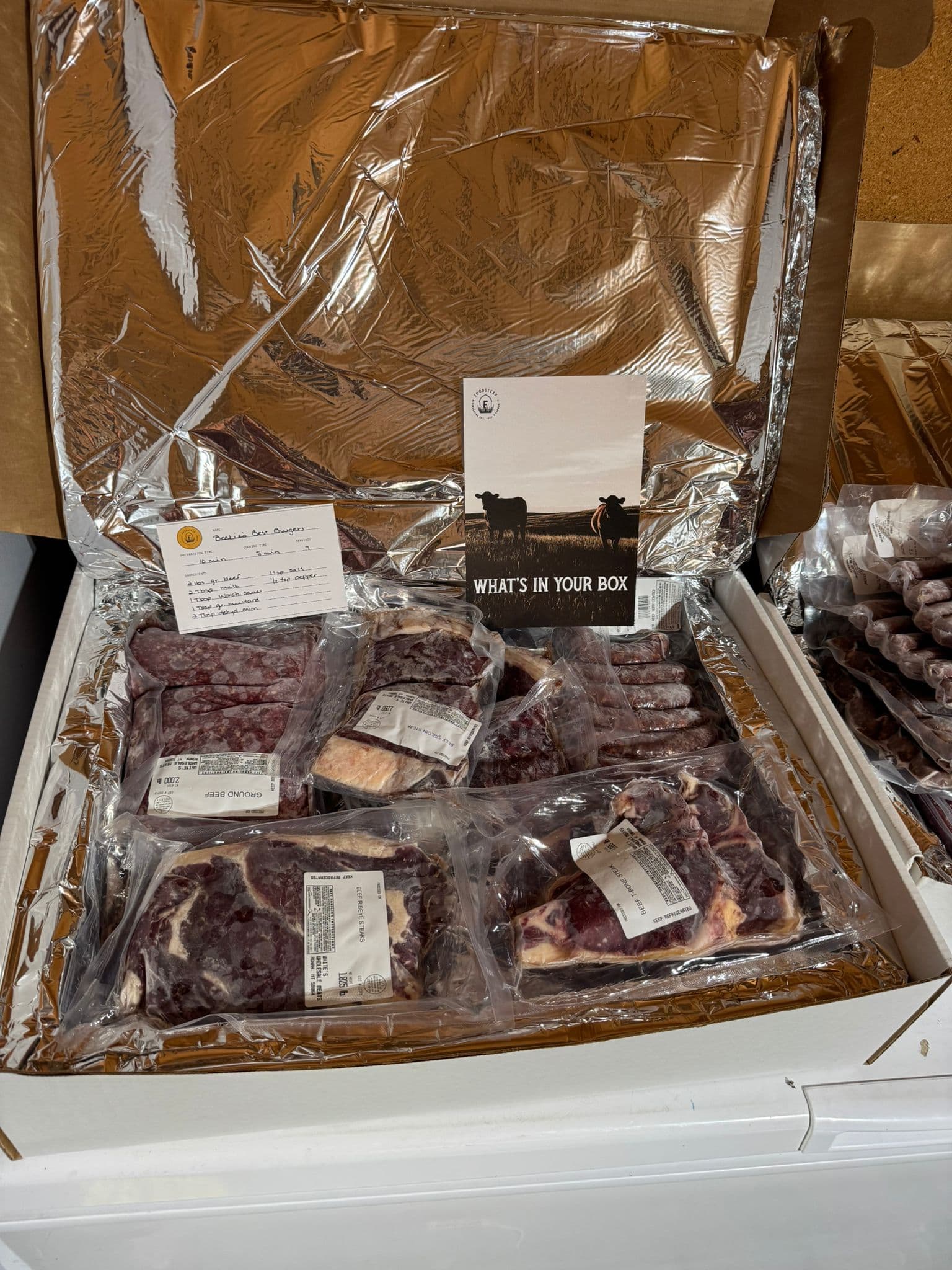 An open Foodstead beef box on a chest freezer, with a What's In Your Box card and Beckie's handwritten recipe card for beef burgers.